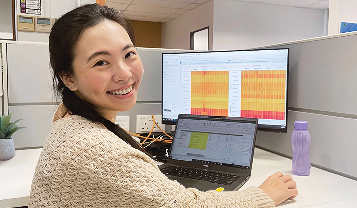 Smiling woman at an office desk with a laptop and monitor displaying colorful heat map data charts.